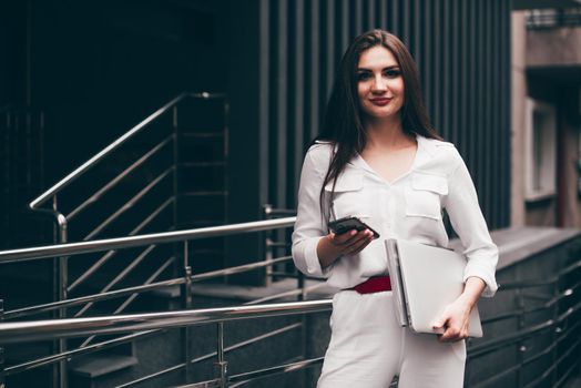 Beautiful young caucasian businesswoman in a white clothes with a laptop and notebook outdoor near business centre.