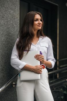 Beautiful young caucasian businesswoman in a white clothes with a laptop and notebook outdoor near business centre.