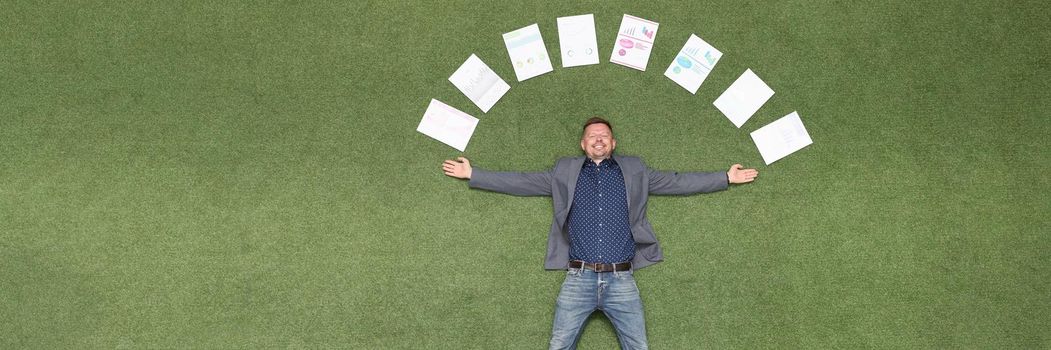 Young businessman lying on green grass with documents around his head top view. Successful business deals concept