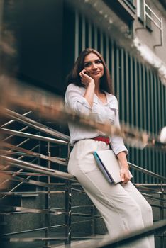 Beautiful young caucasian businesswoman in a white clothes with a laptop and notebook outdoor near business centre.