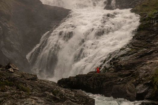 A woman wearing a red dress dancing next to Kjosfossen waterfall in a rocky landscape in Norway