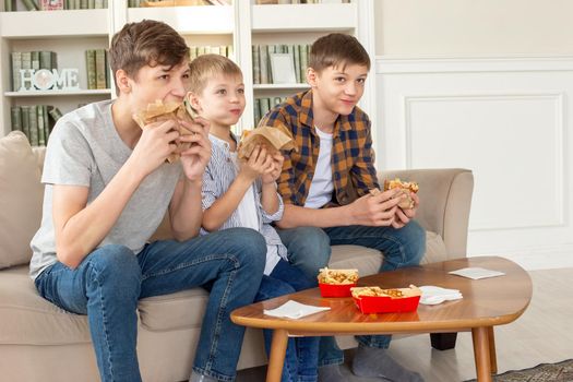 Three cute teenagers, in casual clothes, are sitting on the sofa in the living room, eating fast food, a hamburger, watch tv