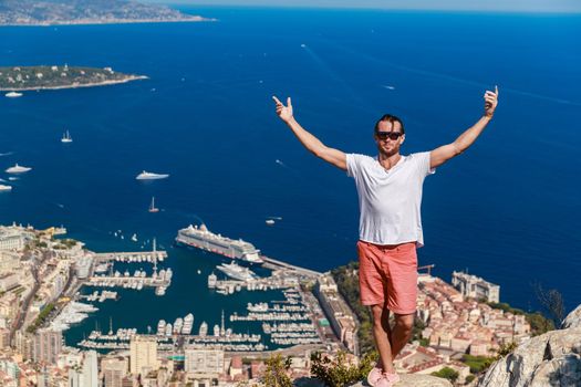 A handsome young man stands and jubilantly with his hands raised on a mountain with the name Of a Head of dog, the Principality of Monaco in the background in clear sunny weather. High quality photo