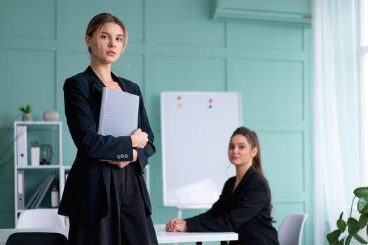 Young women leaders dressed black suit in office one woman standing with document folder other woman sitting on the table looking at camera. Business meeting. Two confidence caucasian female person.