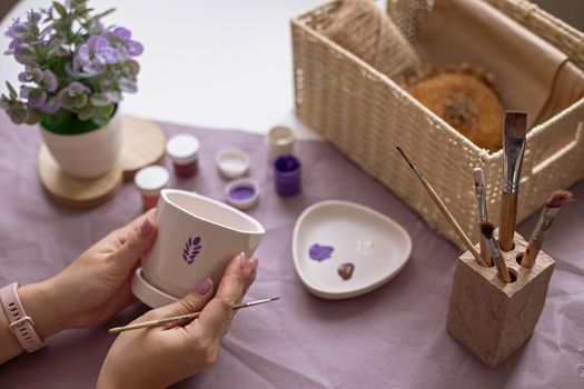 Womens hands with a brush, hold a white ceramic flower pot, with a painted sprig of lavender. On the table are purple craft paper, brushes, paint jars, and a wicker box. Close-up. View from above
