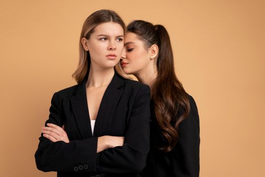 Two young business woman dressed black suit standing studio orange color background. One woman looking looking away arms crossed on chest other woman put her head on her shoulder. Friends support