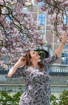 A plump, confident woman stands under a blooming magnolia and smiles. young millennial woman with brown curly hair smiling and waiting for a friend for a date. A full girl enjoys flowering and spring.