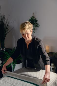 Portrait of a blonde-haired woman, beautician, mature, concentrated and professional, dressed in the black uniform of the company, preparing the massage table to start the day. Relaxing atmosphere and subdued lighting, decoration of plants and candles, background massage table and towels. Vertical