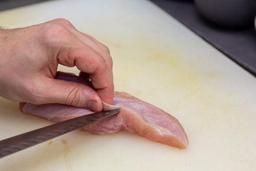 The cook cuts a chicken breast with a knife on a white board.