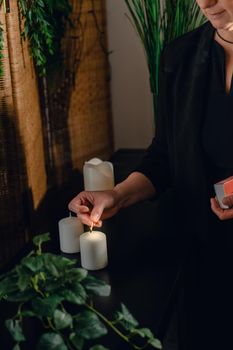 Portrait of a blonde, mature, concentrated and professional beautician, lighting scented candles with a hot wax massage matc.h. Relaxing atmosphere and soft lighting, decoration of plants and candles, massage table in the background and towels. Vertical