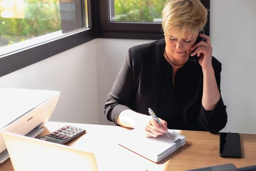 Portrait of a blonde, mature, focused and professional beautician, talking on mobile phone to arrange a visit. She makes a note in the diary of clients' appointments. Owner of a small business Relaxing atmosphere and soft lighting from window, natural light, work table, desk, computer. Horizontal.