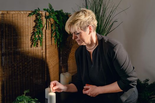 Portrait of a blonde, mature, concentrated and professional beautician, lighting scented candles with a hot wax massage matc.h. Relaxing atmosphere and soft lighting, decoration of plants and candles, massage table in the background and towels. Vertical