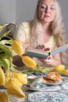 Drinking coffee and tea, a middle-aged blonde woman with long hair drinks tea from a vintage cup and eats puff pastry with pecans on the background of yellow tulips and books on the table