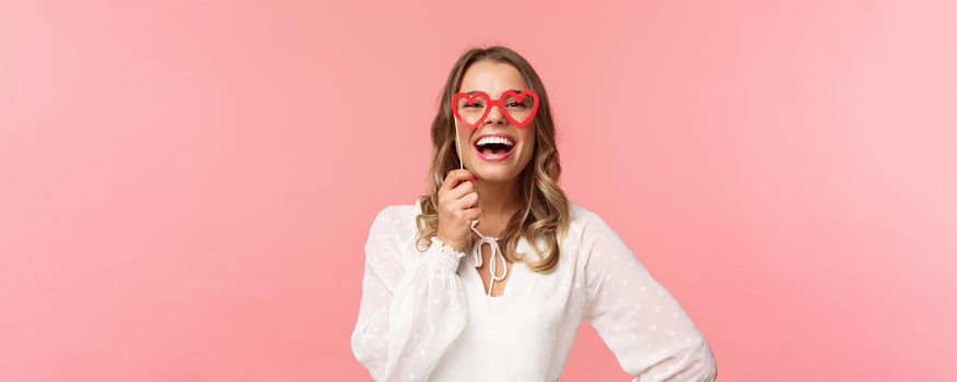 Spring, happiness and celebration concept. Close-up portrait of funny and carefree, beautiful caucasian woman with blond hair, white dress, holding heart-shaped glasses mask and laughing.