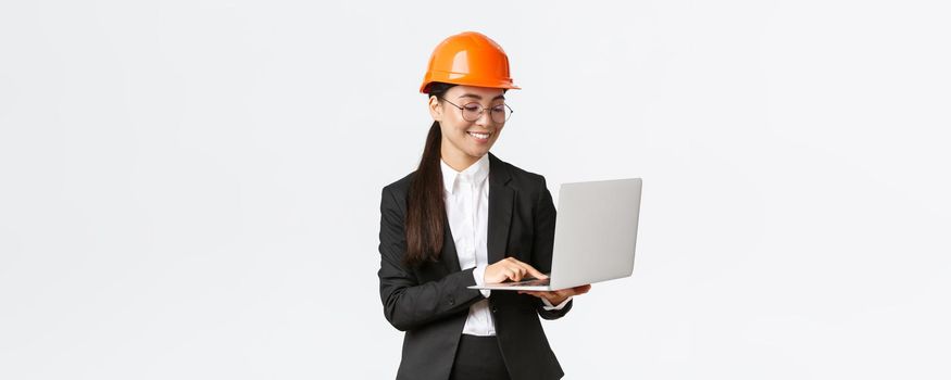 Smiling successful female asian industrial engineer, factory manager in safety helmet and business suit using laptop computer, checking with project or blueprints on screen.