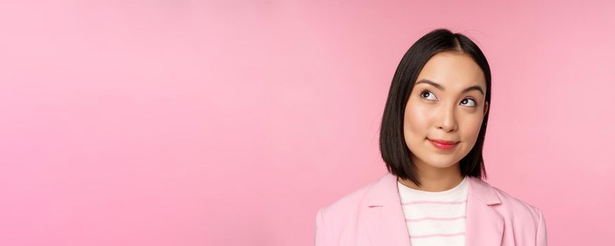 Close up portrait of young asian businesswoman thinking, smiling thoughtful and looking at upper left corner, standing over pink background.