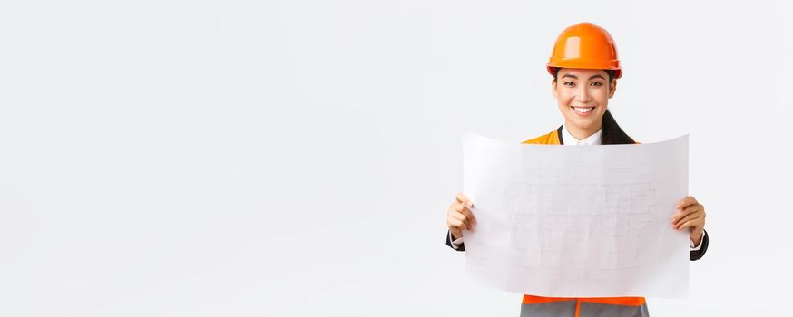 Excited smiling asian female engineer at construction area study blueprints, looking pleased while reading documents, building plan, standing in safety helmet over white background.