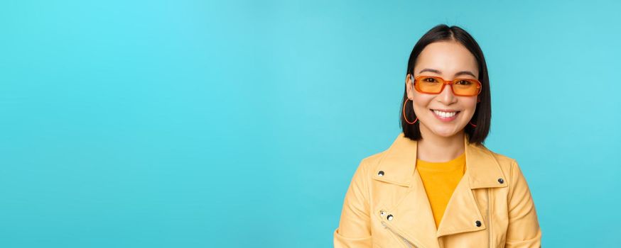 Close up portrait of smiling stylish asian woman in sunglasses, looking happy at camera, posing in trendy yellow jacket, blue background.