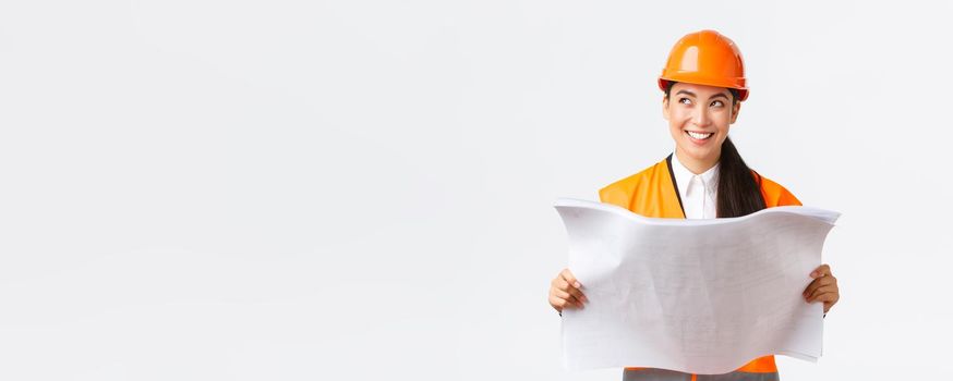 Creative asian female architect imaging her plan, standing in safety helmet and looking away thoughtful while reading blueprints, picturing buiding after finish construction works, white background.