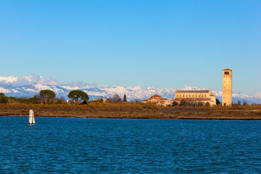 The coastline and high bell tower on the island of Burano with the snowy mountains behind