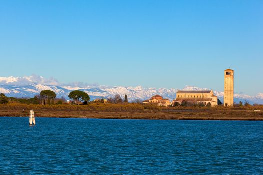 The coastline and high bell tower on the island of Burano with the snowy mountains behind