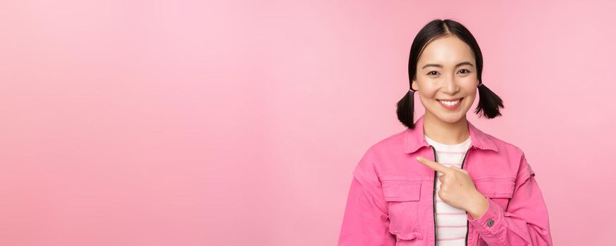 Portrait of beautiful smiling asian girl, pointing finger left, showing advertisement, banner promo, standing over pink background.