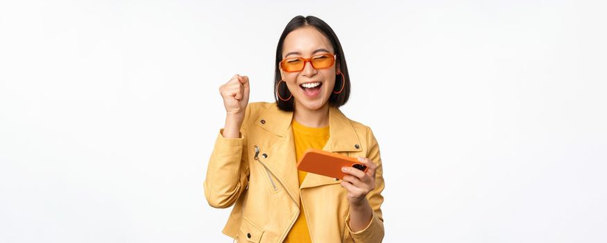 Image of stylish korean girl dancing with smartphone, laughing happy and smiling, standing over white background. Copy space