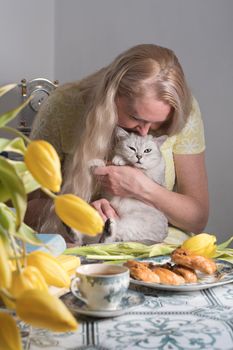 blonde middle-aged hugging a kitten and drinking tea, still life with yellow tulips and cakes a stack of books on the table. High quality photo