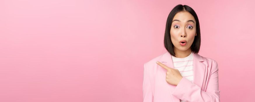 Portrait of businesswoman with surprised face, pointing finger left, showing smth interesting, standing over pink background in office suit.
