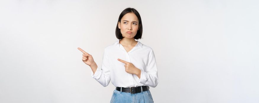 Disappointed, sad young asian woman pointing and looking left with upset, sulking face expression, standing over white background.