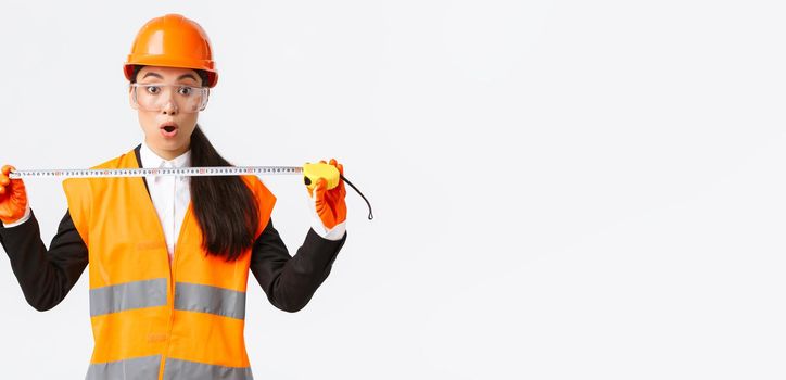 Surprised and excited asian female construction engineer, industrial worker in safety helmet and uniform, measuring layout, holding tape measure and stare amazed at camera, white background.