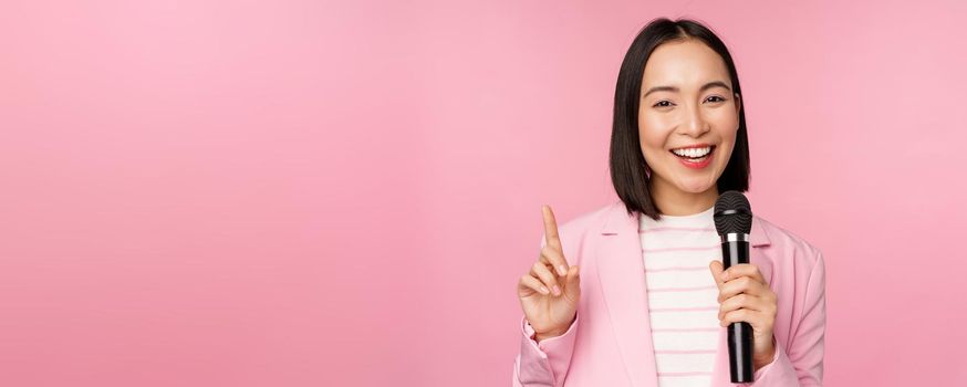 Image of enthusiastic asian businesswoman giving speech, talking with microphone, holding mic, standing in suit against pink studio background.