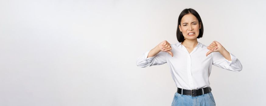 Dislike. Disgusted asian woman showing thumbs down and grimacing, commenting on smth bad, disagree, complaining, standing over white background.