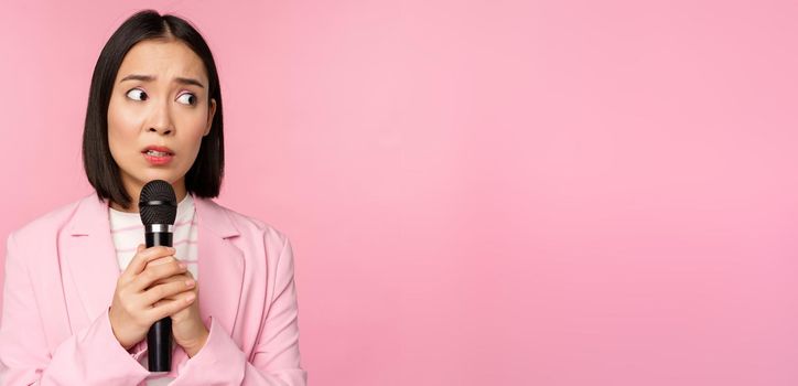 Insecure asian businesswoman giving speech, scared of talking in public, using microphone, standing in suit over pink background.
