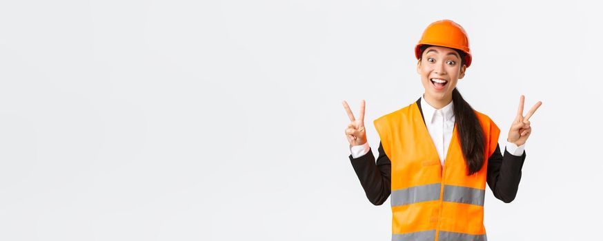 Happy smiling asian female engineer, architect in safety clothins and helmet, showing peace kawaii sign and looking upbeat, company winning tender on construction and building, white background.