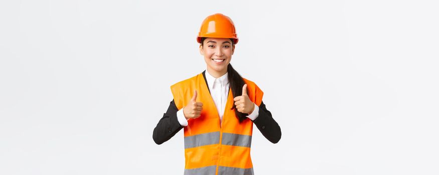 Upbeat pleased asian female architect giving permission, proud with result, standing in safety helmet and reflective jacket, showing thumbs-up in approval, guarantee best construction quality.