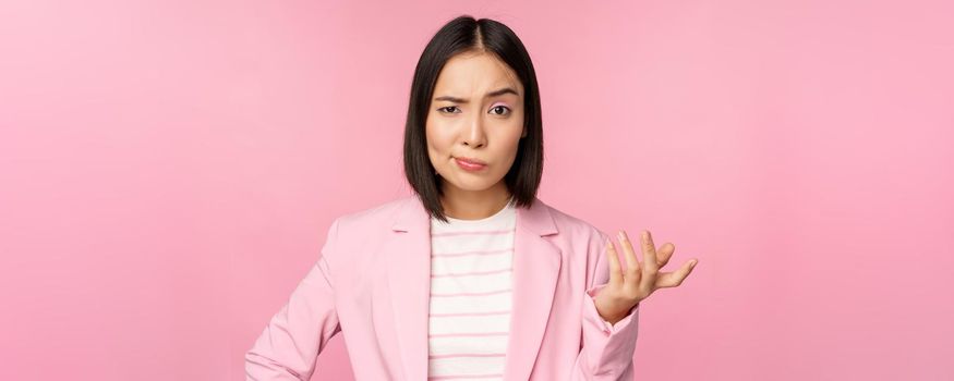 Portrait of angry asian woman in suit, clench fists and looking furious, outraged of smth bad, standing over pink background.