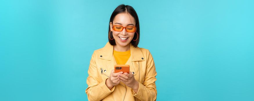 Smiling asian girl in sunglasses, using smartphone app, holding mobile phone, standing over blue background. Copy space