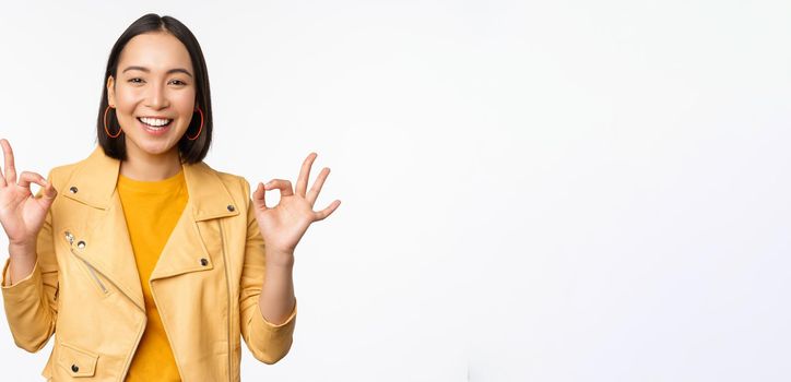 Happy beautiful asian girl shows okay, ok recommending sign, pleased by smth, praising quality, standing over white background. Copy space