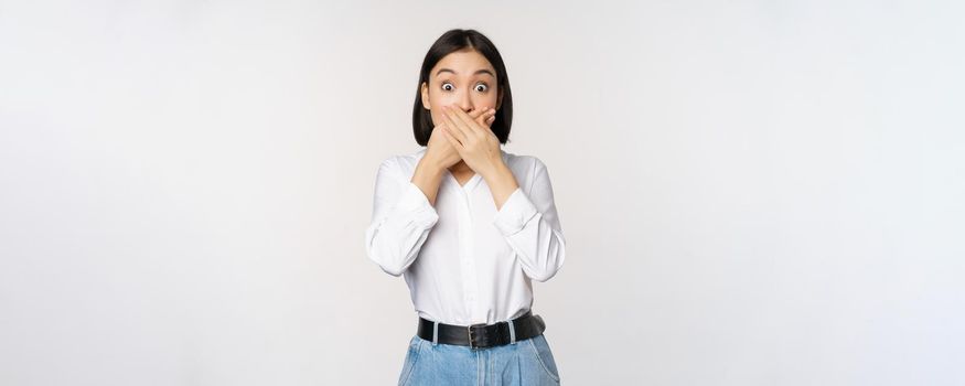 Shocked young asian girl close mouth, cover lips with hands and looking amazed, standing over white background. Taboo gesture