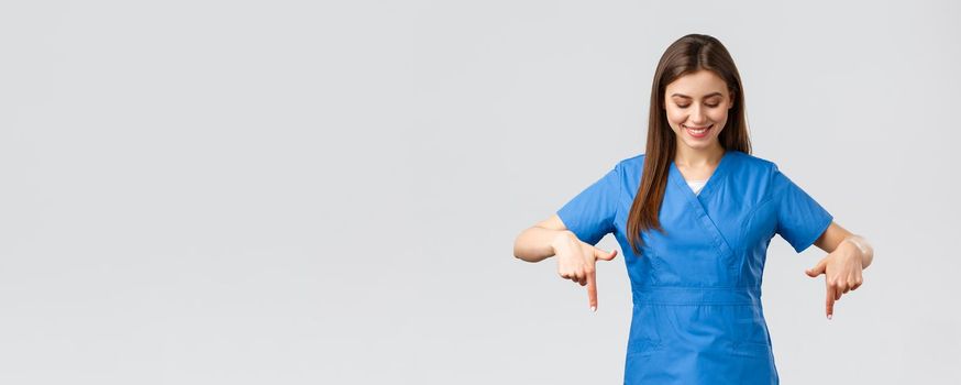 Healthcare workers, prevent virus, covid-19 test screening, medicine concept. Happy and pleased attractive female nurse or doctor, physician in blue scrubs, pointing looking down at advertisement.