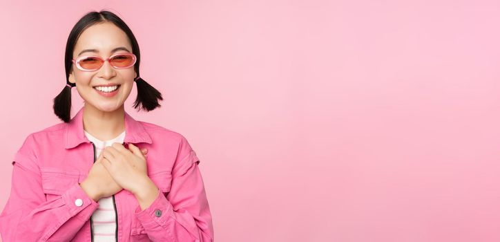 Smiling young asian woman in sunglasses looking thankful, holding hands on heart, flattered, appreciate smth, standing over pink background.
