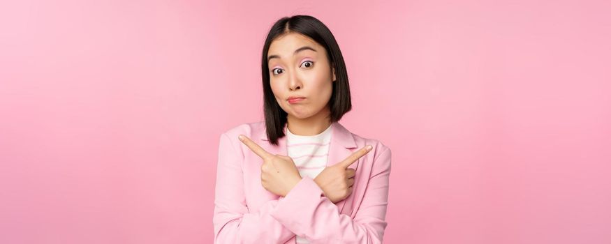 Decision concept. Young asian businesswoman, corporate worker pointing fingers sidways, two directions, showing variants, looking indecisive while choosing, pink background.