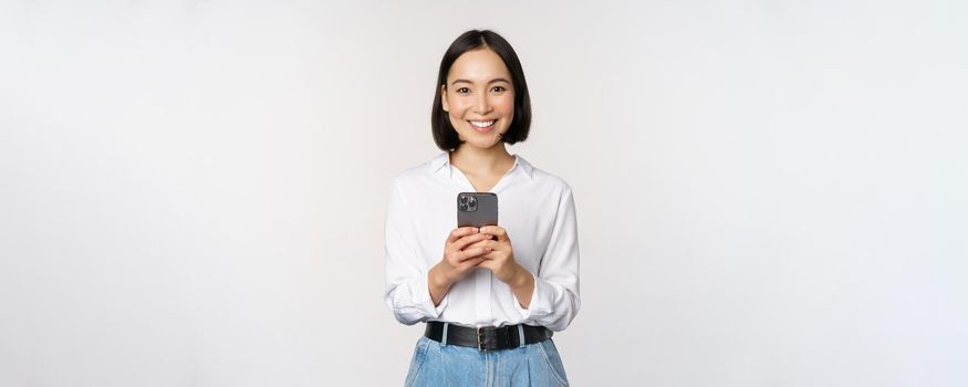 Stylish modern asian girl using mobile phone application, chatting on cellphone and smiling, standing in white blouse against studio background.