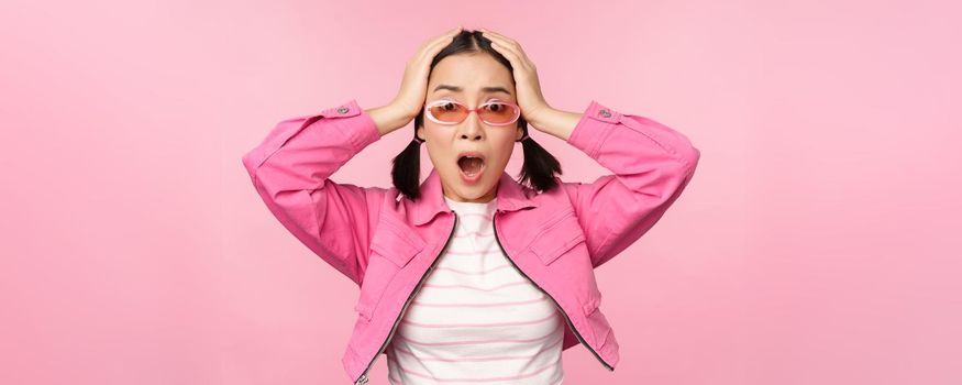 Portrait of troubled asian woman, korean girl holds hands on head and look in panic, standing distressed and shocked against pink background.