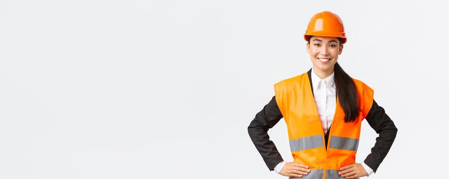 Successful young female asian businesswoman, architect in safety helmet, reflective jacket, standing confident and determined, smiling assured, building houses, posing over white background.