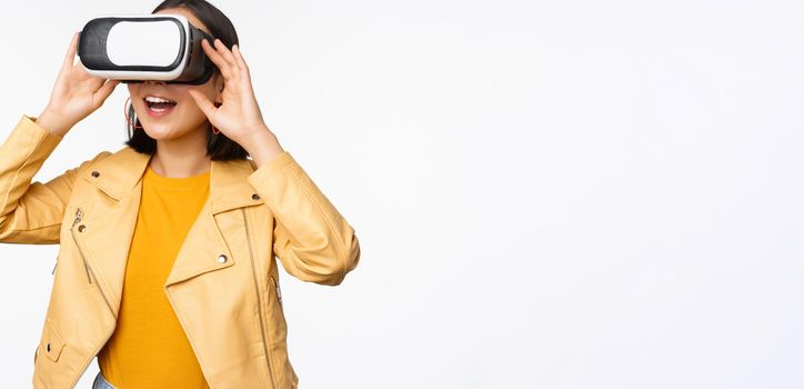 Enthusiastic asian girl in VR glasses, looking with virtual reality headset at smth amazing, standing over white background. Copy space