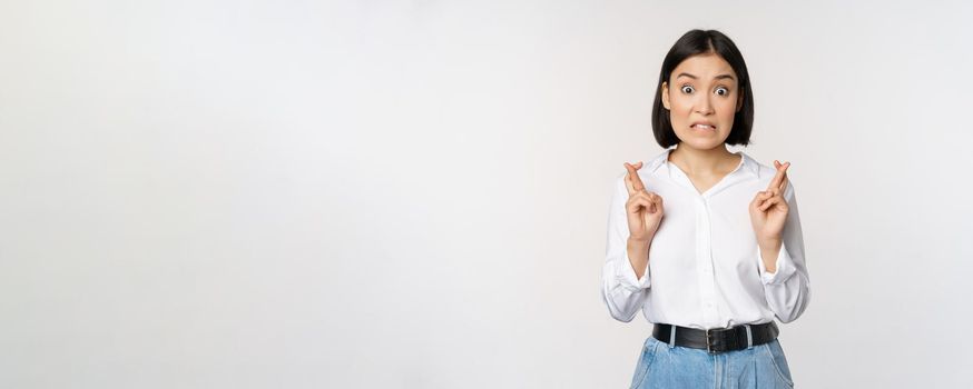 Portrait of young korean woman, asian girl cross fingers and praying, making wish, anticipating, waiting for results, standing over white background.