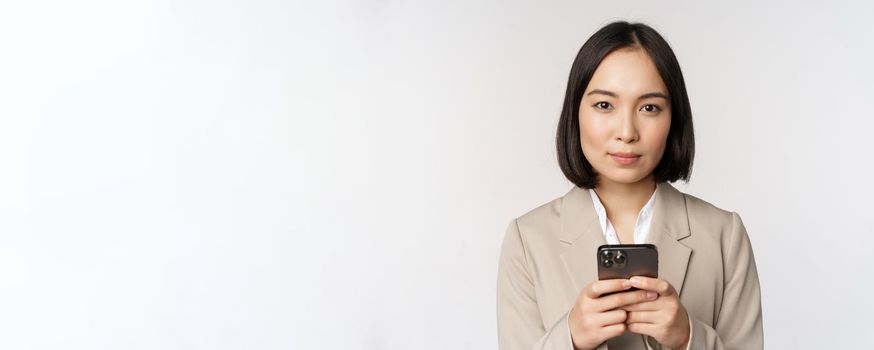 Image of asian businesswoman in suit, holding mobile phone, using smartphone app, smiling at camera, white background.