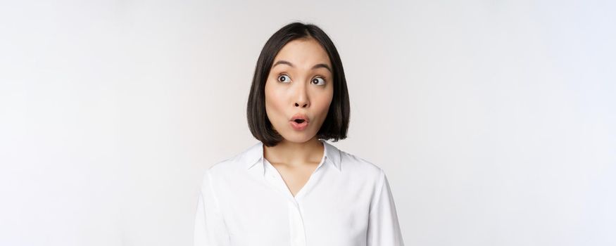 Close up portrait of young asian woman making surprised face, looking at upper left corner impressed, wow emotion, standing over white background.